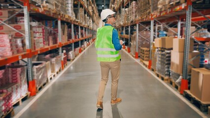 Caucasian man in helmet tapping on tablet while walking in warehouse of modern factory. Back view. Middle-aged male worker doing inventory in construccion store using digital device, retail storehouse