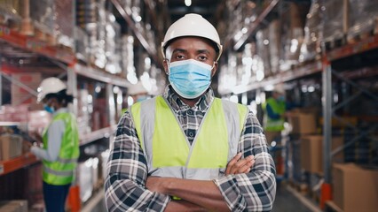 African American male worker standing in hard hat and face mask in retail warehouse with many shelves with boxes and goods looking at camera. Business, coronavirus, logistics center, close up