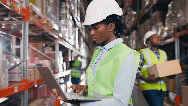 Professional African American female worker wearing hard hat uses laptop typing checking inventory standing in retail warehouse with shelves. Working in logistics, distribution center, job concept