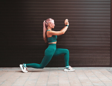 Fitness smiling woman in grey sports clothing with pink hair. Young beautiful model with perfect body.Female posing in the street near  wall.Doing forward lunges. Stretching out before training