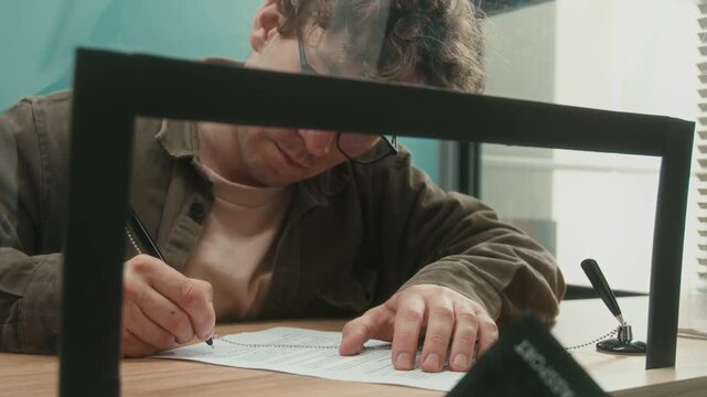 Shot from behind service window glass of male client signing document to withdraw money then receiving cash from bank teller at counter