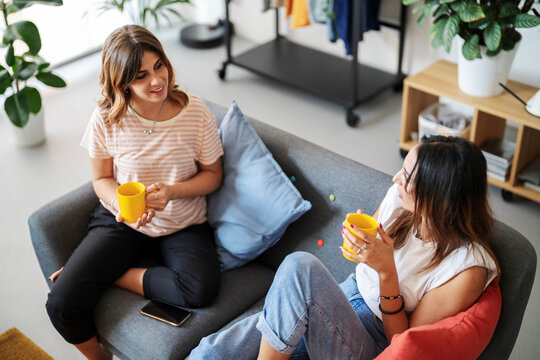 Two young women are comfortably seated on a modern sofa, each holding a yellow mug. They are engaged in a pleasant conversation, sharing a relaxed moment in a bright living space.