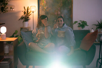 Two women enjoy a cozy movie night at home. One covers her face in fear while the other points at the screen, both eating popcorn. They are immersed in the film.