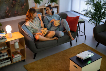 Two young women relax on a sofa, eating popcorn and chatting. A projector is set up, ready for a movie night or home entertainment. They enjoy a cozy evening together.