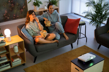 Two young women are relaxing on a sofa at home, enjoying a movie night. They are eating popcorn and watching a film projected onto a screen.