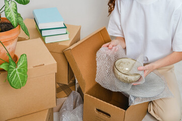 Unrecognized woman wearing white t-shirt sitting on the floor surrounded with carton boxes and packing plates and dishes. Relocating, buying new house. Pack personal stuff from carton boxes. 
