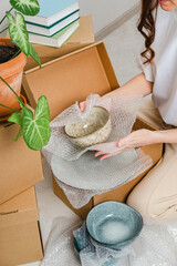 Unrecognized woman wearing white t-shirt sitting on the floor surrounded with carton boxes and packing plates and dishes. Relocating, buying new house. Pack personal stuff from carton boxes. 