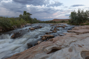 The shallow Virgin River in Southern Utah USA rushes over sandstone ledges and boulders next to rain filled potholes in the rocks under a cloudy evening sky with a touch of pink light on the horizon.