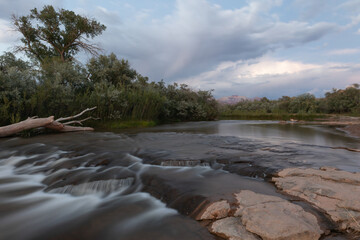 The shallow Virgin River lined by cottonwood and willow trees, pours over sandstone ledges under a sky filled with the remnants of a summer monsoon storm.