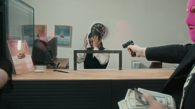 Medium shot of masked female robber robbing bank with male accomplice pointing gun at scared cashier behind tellers counter raising hands up then dialing 911 with shaking hands