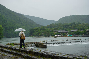 Lone man holding an umbrella in the rain looks towards the forests and mountains surrounding Arashiyama, Kyoto, Japan, on the Katsura River next to the Togetsukyo Bridge