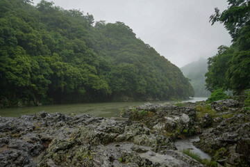 The Katsura River flows through a forest and hilly landscape surrounded by fog in the rain in Arashiyama, Kyoto, Japan