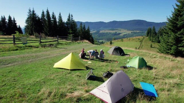 Aerial view campsite in mountains, with several colorful tents on grassy clearing. Campers relax under clear blue sky, surrounded by tall evergreens and panoramic views of distant peaks.