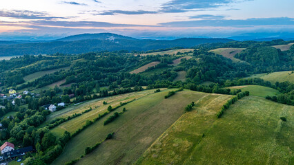 Summer aerial view of the Bieszczady Mountains – rolling fields, forested hills, and rural buildings. Picturesque landscape at sunset.