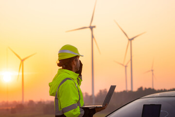 Engineer working on laptop in front of wind turbines for checking wind turbines of the field during beautiful sunset. Alternative energy concept.