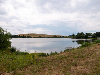 Calm fishing pond with grassy shore and distant fields. Peaceful rural landscape near Vrbov village, Slovakia.
