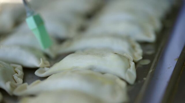 Tight close-up clip captures raw empanadas being meticulously brushed with oil before oven baking. Focus on glistening texture and preparation ritual.