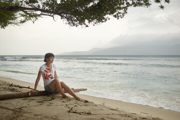 A contemplative woman sits on a piece of driftwood on a quiet, sandy beach, gazing at the ocean...
