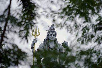 lord shiva behind green leaves, shiva statue through trees, peaceful shiva nature view, majestic deity surrounded by nature,spiritual god in jungle frame, nature framed shiva sculpture.





