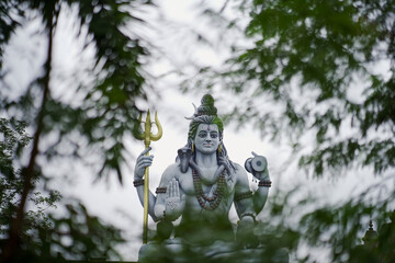 lord shiva behind green leaves, shiva statue through trees, peaceful shiva nature view, majestic deity surrounded by nature,spiritual god in jungle frame, nature framed shiva sculpture.





