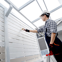 Fototapeta premium Asian man painter standing on the ladder painting white acrylic color on house wooden wall. Male decorator using paintbrush for home renovation.