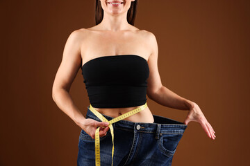 Fit woman with measuring tape wearing old jeans after weight loss on brown background, closeup