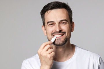 Smiling man applying lip balm onto his lips on grey background
