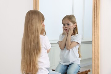 Upset little girl looking at herself in mirror indoors