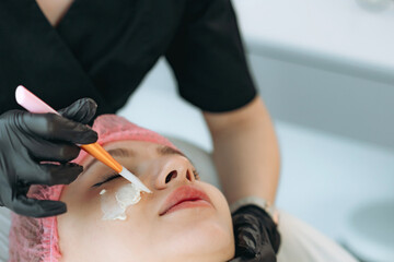 professional cosmetologist applies a creamy face mask on yiung woman face using a brush