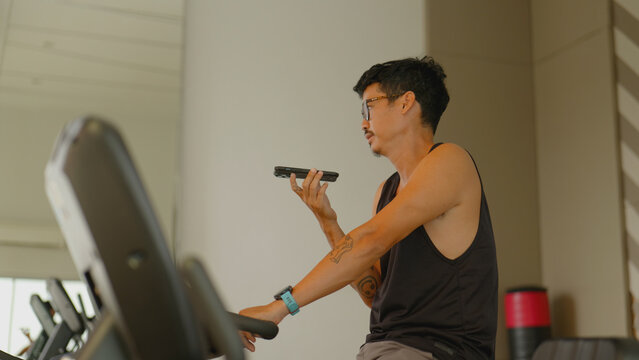 Asian man rides a stationary bike at the gym while talking on his smartphone.