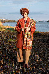 Woman in an orange coat and plaid scarf stands by a burnt field near water