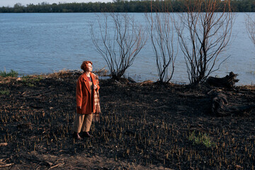 Woman in an orange coat stands on a burnt field by a serene lake