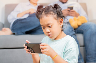 Little daughter sit on floor playing game on smartphone while her parent sitting on sofa also using their mobile phone.