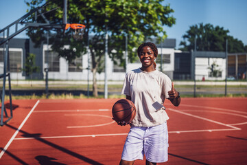 Naklejka premium Young African American woman standing on sunny urban basketball court, holding a ball and giving thumb up with cheerful smile, representing joy and active lifestyle.