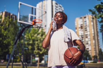 Young black woman is standing on basketball court. She is tired and wiping sweat of her face.