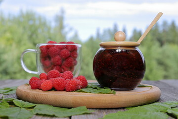 A wooden plate with a glass jar of fresh raspberry jam, heap of berries and a glass mug of ripe raspberries is placed among raspberry leaves on an old wooden table  against a summer forest landscape. 