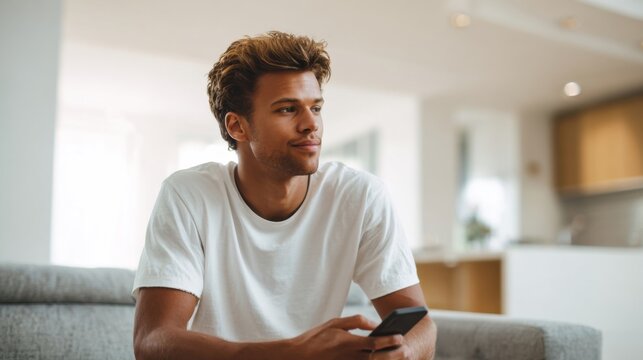 Young man sitting on sofa indoors using smartphone with modern kitchen background.