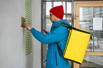 Courier with a bright yellow thermal backpack ringing the doorbell while delivering a food order at...