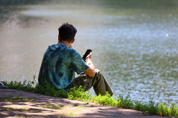 Guy sitting with smartphone in hand on lake beach. Mobile communication in summer park