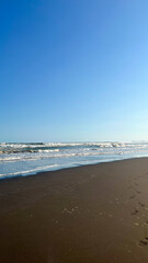 A peaceful beach scene with gentle waves meeting the sandy shore beneath a clear blue sky. The calm ocean and expansive sky create a serene coastal atmosphere.