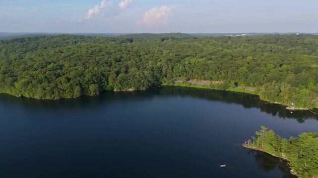Calm and deep blue water of Kensico Reservoir, vast green forest and scenic road surround lake, Westchester County, New York, USA. Aerial drone lateral view
