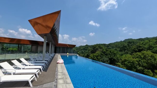 A beautiful time-lapse shows fluffy clouds moving over a stunning empty infinity pool, modern architectural canopy, and lounge chairs at a luxury hotel nestled in a lush green forest - low demand