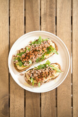 Two open faced sandwiches featuring toasted rye bread, creamy hummus, fresh arugula, and spicy sausage crumbles, presented on a white plate atop a wooden table