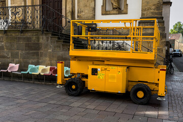 Yellow lift parked beside colorful chairs on street.