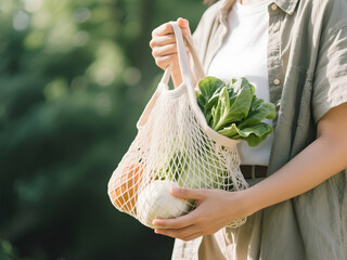 Woman holding cream mesh bag filled with leafy greens and vegetables in soft natural garden setting with sunlit greenery in background