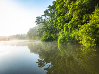 Morning landscape: lake in fog with reflection of green forest under blue sky.