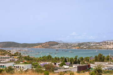 A scenic view of a coastal town with boats in the harbor. The landscape features hills, greenery,...