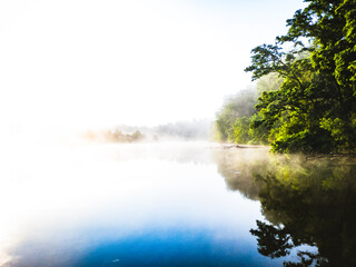 Morning landscape: lake in fog with reflection of forest under blue sky.