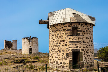 Three historic windmills stand on a hillside under a clear blue sky. The structures are made of...