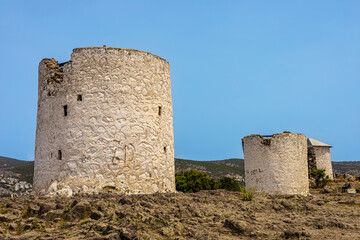 Three ancient stone windmills stand on a rocky landscape under a clear blue sky. The structures are weathered and show signs of age.